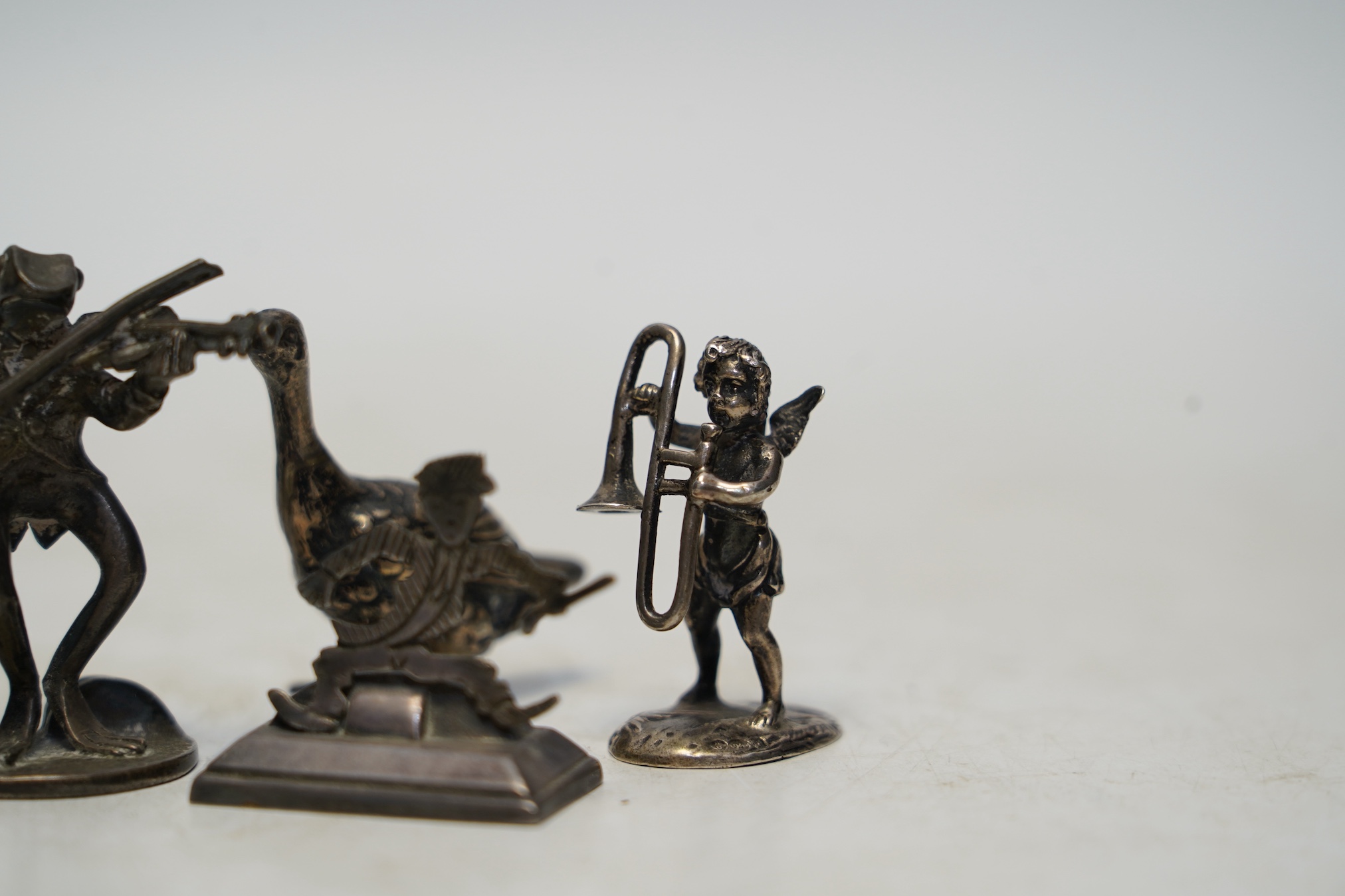 Two early 20th century miniature silver models of a goose and a cherub with trombone, 37mm, an 800 standard figural menu holder and a later silver model of a frog with violin. Condition - fair